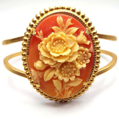 Front view of the retro vintage floral cameo hinged bangle bracelet. The metal is gold tone in color. The front has a large oval with three flowers in the middle in yellow tones. The background of the plastic cameo is shades of orange.