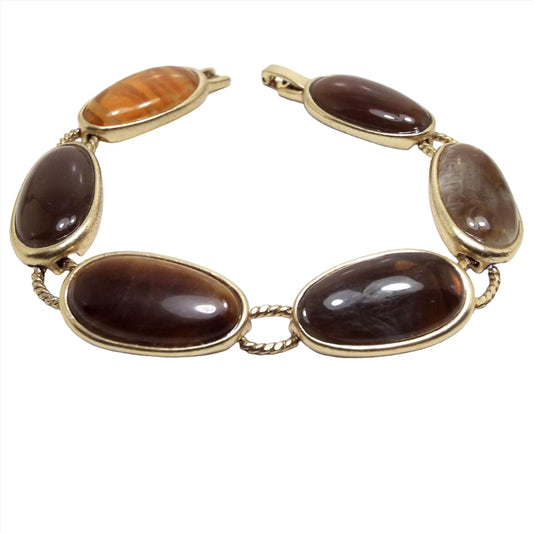 Angled top view of the retro vintage Liz Claiborne link bracelet. The metal is gold tone in color. The links are oval thin egg like shaped. Most are brown with a couple that have shades of orange and yellow. The fourth one from the clasp has a tiger's eye gemstone cab. The rest are resin.
