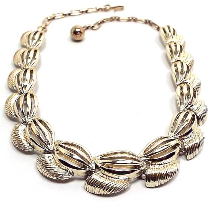 Front view of the retro vintage gold tone metal link choker necklace. The links have a football shape oval design with cut out curvy lines on them. At the bottom of the links is a flared corrugated area. There is a chain and hook clasp at the end.