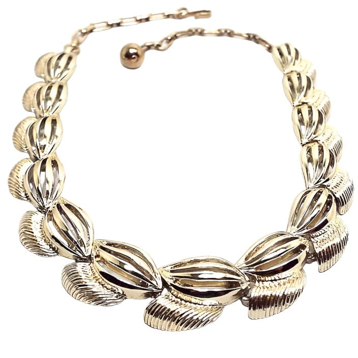 Front view of the retro vintage gold tone metal link choker necklace. The links have a football shape oval design with cut out curvy lines on them. At the bottom of the links is a flared corrugated area. There is a chain and hook clasp at the end.
