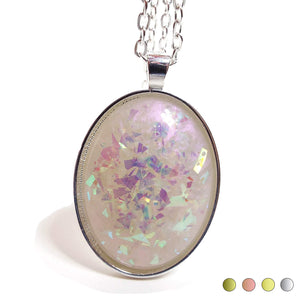 Front view of the large oval handmade resin pendant. The oval resin cab is pearly color shift with light hues of off white and pink. There are larger pieces of AB glitter on the inside of the resin with flashes of pink, purple, blue, and green. There are four color swatches on the bottom right showing it comes in a choice of antiqued brass, rose gold tone, gold tone, and silver tone plated.