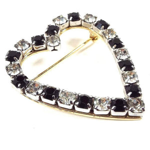 Angled front view of the retro vintage rhinestone heart brooch pin. the metal is gold tone in color. The middle area of the heart is open. There are small round rhinestones alternating in color in clear and black all the way around the brooch.