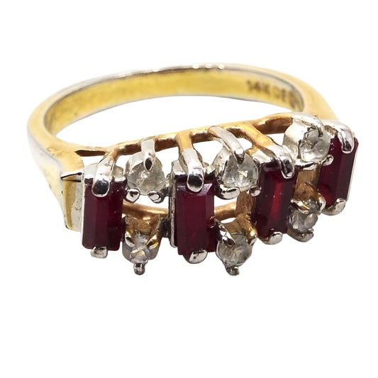 Front view of the retro vintage rhinestone cocktail ring. The metal is gold tone in color with some rub wear on the band showing the silver tone underneath. There are four rectangle baguette dark red rhinestones on the top with two small round clear rhinestones in between each red one at the top and bottom. All stones are prong set.