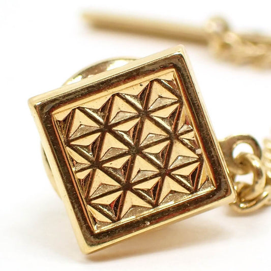 Enlarged front view of the retro vintage geometric tie tack. It is square shaped with gold tone plated color metal. There is a raised triangle pattern all across the front.
