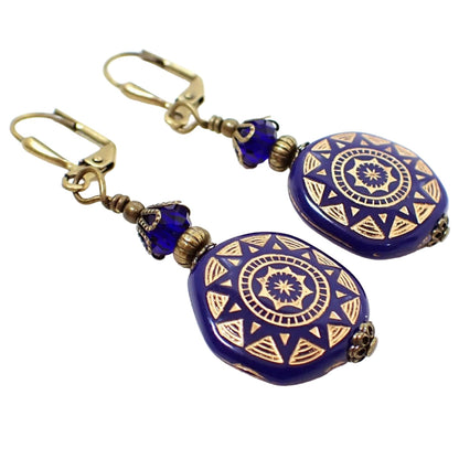 Angled view of the handmade coin shape drop earrings. The metal is antiqued brass in color. There are cobalt blue faceted glass crystal beads at the top. The bottom beads are coin shaped with a metallic sun design painted on them.