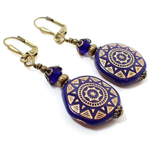 Angled view of the handmade coin shape drop earrings. The metal is antiqued brass in color. There are cobalt blue faceted glass crystal beads at the top. The bottom beads are coin shaped with a metallic sun design painted on them.