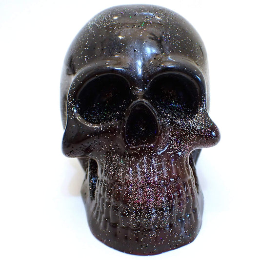 Front view of the handmade resin skull. It is primarily pearly black in color with some purple around the teeth and jaw area. There is silver holographic glitter scattered around for sparkle and tiny flashes of color.