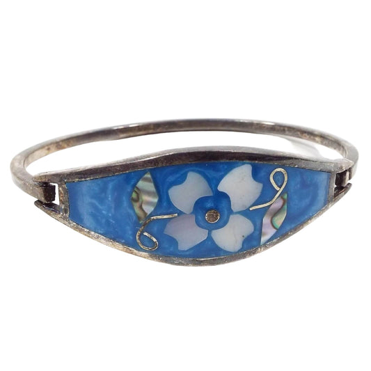 Front view of the retro vintage Southwestern hinged bangle bracelet. The metal is silver tone in color. The front is pearly blue enameled with a floral design in the middle. The flower petals are inlaid mother of pearl shell and the leaves are inlaid abalone.