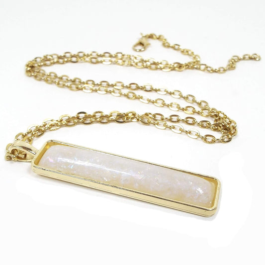 Angled view of the AB sparkle resin handmade pendant necklace. The metal is gold tone in color. There is a cable link chain with lobster clasp at the end. The pendant is a long rectangle bar shape with a domed pearly off white resin cab that has AB sparkles and flashes of color in it..