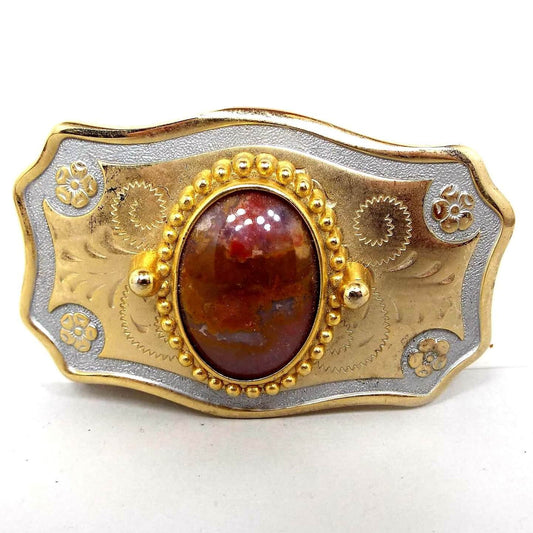 Front view of the retro vintage agate gemstone belt buckle. The buckle is mostly gold tone in color with silver tone around the edge. There are flowers at the corners and an etched design in the middle area. In the very middle is an oval agate cab with shades of brown and orange.