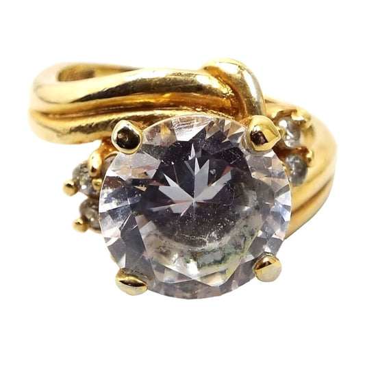 Front view of the retro vintage rhinestone cocktail ring. The metal is gold tone in color. the band curves around each part of the top part. There is a round clear rhinestone in the middle and a cluster of three smaller round clear rhinestones on each side. All are prong set.