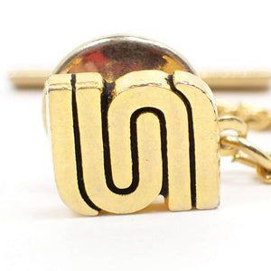 Enlarged front view of the retro vintage tie tack. The metal is gold tone plated in color. There are curvy U like shapes fitted together in opposite directions with curved areas on the sides. The inside areas of these shapes have black paint for definition.