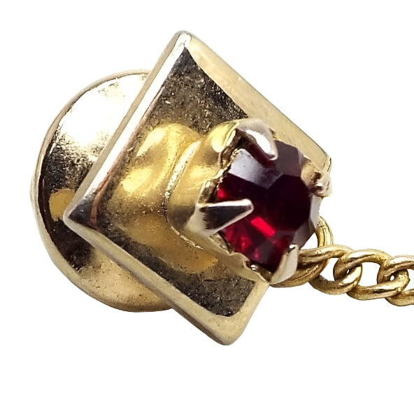 Enlarged front view of the Mid Century vintage rhinestone tie tack. The metal is gold tone in color. It's square in shape with a small round red rhinestone in the middle.