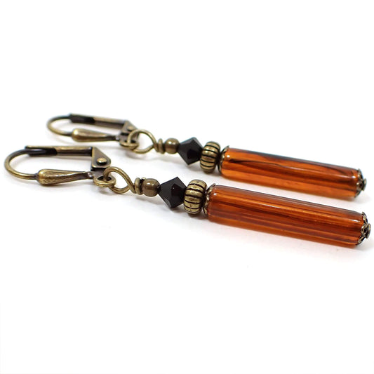 Side view of the handmade faux tortoise stick earrings. The metal is antiqued brass in color. There are faceted black glass crystal beads at the top. The bottom lucite beads are tube shaped stick like and have shades of orange, brown, and black in them. 