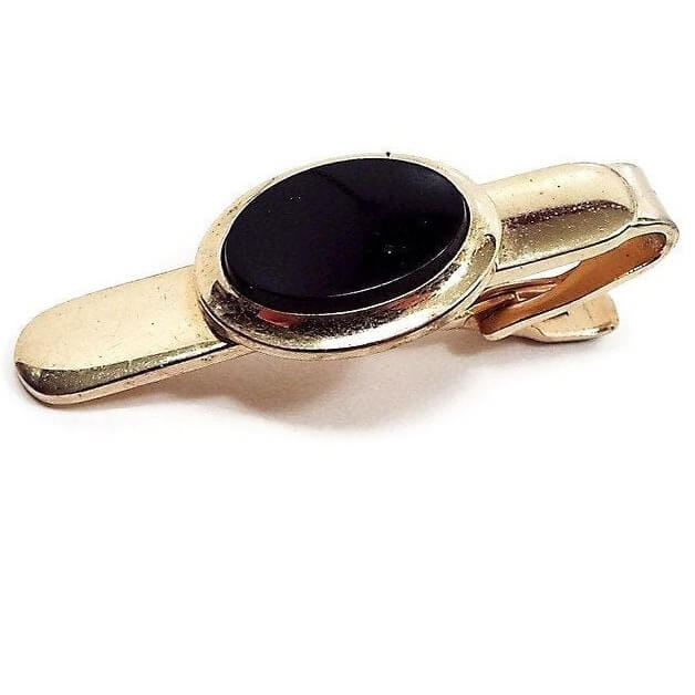 Front view of the retro vintage Speidel tie clip. The metal is gold tone in color. There is a black oval plastic cab in the middle. 