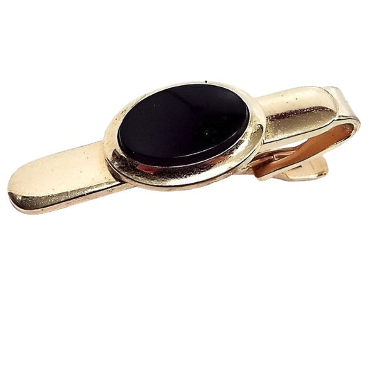 Front view of the retro vintage Speidel tie clip. The metal is gold tone in color. There is a black oval plastic cab in the middle. 