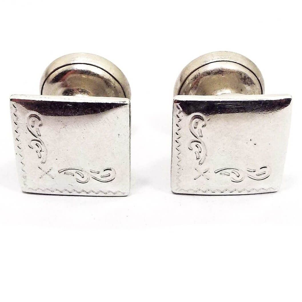 Front view of the 1930's vintage cufflinks. They are silver tone in color. The fronts have squares with a lightly etched pattern on the edge of one side and the bottom. There is a round part on the back that pulls out and retracts with a chain.