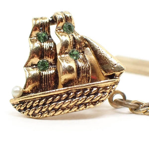 Enlarged front view of the Mid Century vintage ship tie tack. The metal is gold tone in color. It is shaped like an old ship with sails. The sails have green rhinestones on them and there is a faux pearl at the front of the boat.