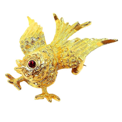 Front view of the Mid Century vintage Mamselle brooch pin. The metal is gold tone in color. It is shaped like a small bird with its feet out and its wings spread towards the tail. There is a red rhinestone for the eye.