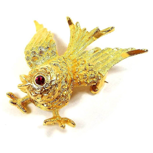 Front view of the Mid Century vintage Mamselle brooch pin. The metal is gold tone in color. It is shaped like a small bird with its feet out and its wings spread towards the tail. There is a red rhinestone for the eye.