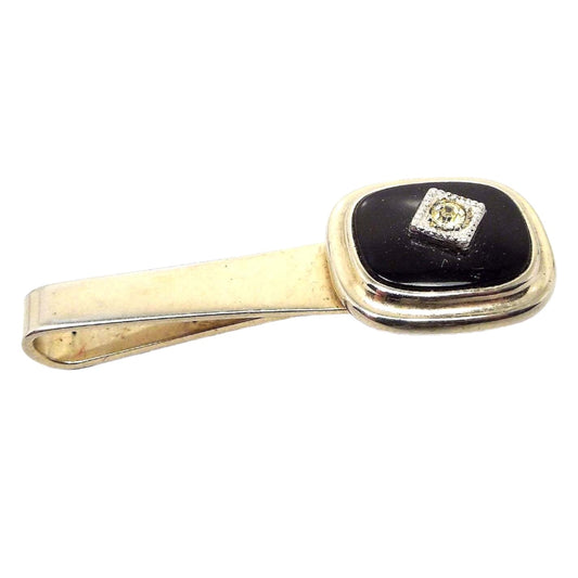 Angled front view of the Mid Century vintage slide on tie bar. The metal is gold tone in color. At the end is a rounded square of black glass with a rhinestone in the middle.