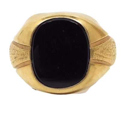 Front view of the retro vintage Uncas onyx ring. The metal is gold tone in color. It is styled like an old signet ring with a rounded rectangle black onyx cab in the middle and flared out sides. There are engraved lines on each side with a textured metal part in between.