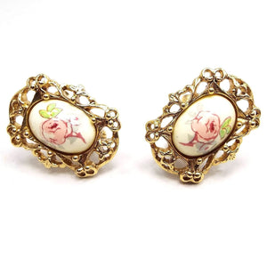 Front view of the retro vintage floral clip on earrings. The metal is gold tone in color and there is a filigree setting around a plastic oval cab in the middle. The cabs have decals with pink rose flowers on them. 
