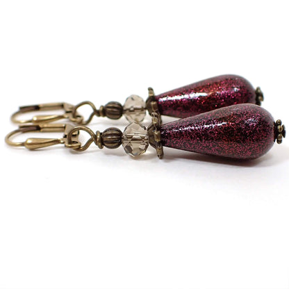 Side view of the handmade glitter drop earrings. The metal is antiqued brass in color. There are faceted glass crystal beads at the top in a light brownish gray color. The bottom acrylic beads are teardrop shaped and are black and coated with fine magenta purple red glitter with a few specks of red mingled in. 