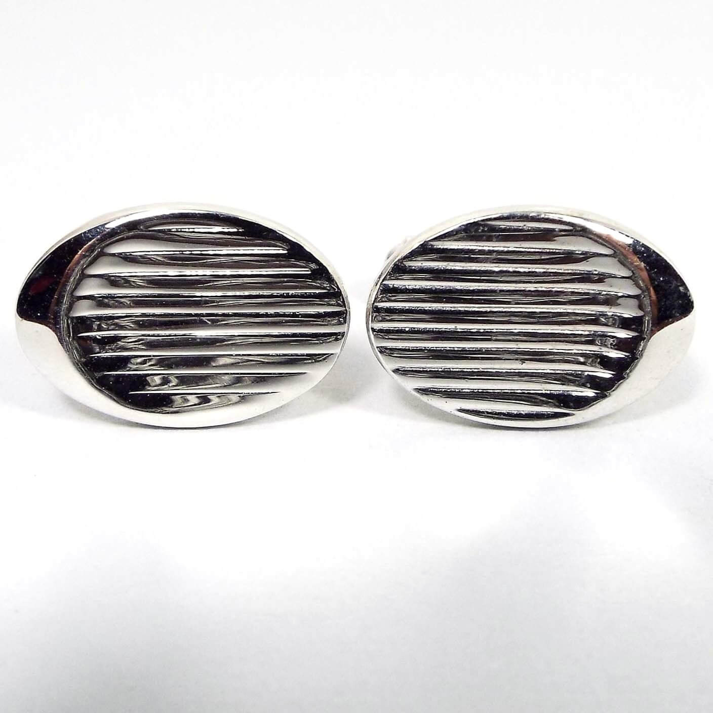 Front view of the Mid Century vintage Modernist style Emmons cufflinks. They are oval in shape and have a line design. The shiny silver color metal is picking up dark reflections from the background.
