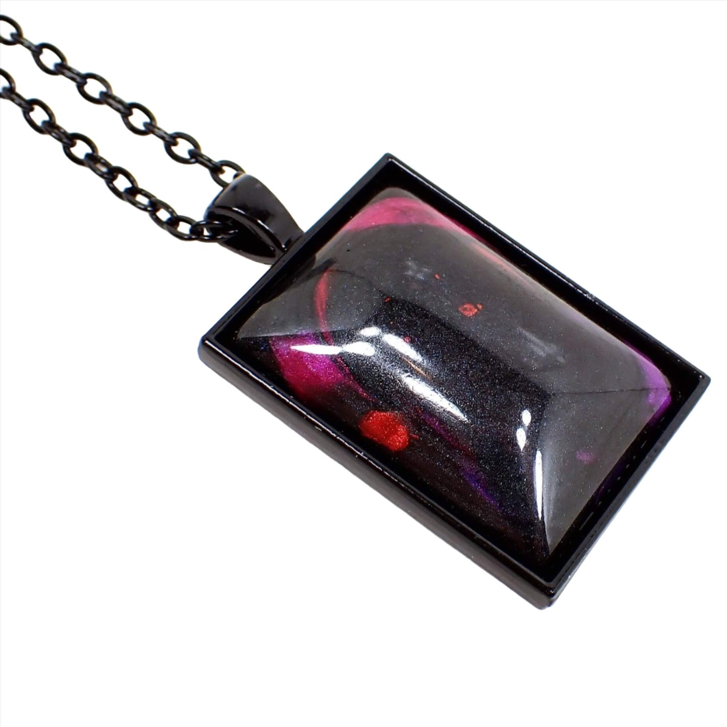 Angled enlarged view of the handmade Goth large rectangle pendant necklace. The chain and setting are black in color. There is a large rectangle pendant with a pearly black domed resin cab. There are splashes of bright pearly pink, red, purple, and blue here and there on the pendant.