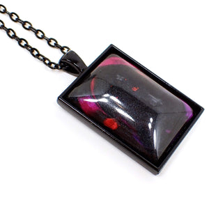 Angled enlarged view of the handmade Goth large rectangle pendant necklace. The chain and setting are black in color. There is a large rectangle pendant with a pearly black domed resin cab. There are splashes of bright pearly pink, red, purple, and blue here and there on the pendant.
