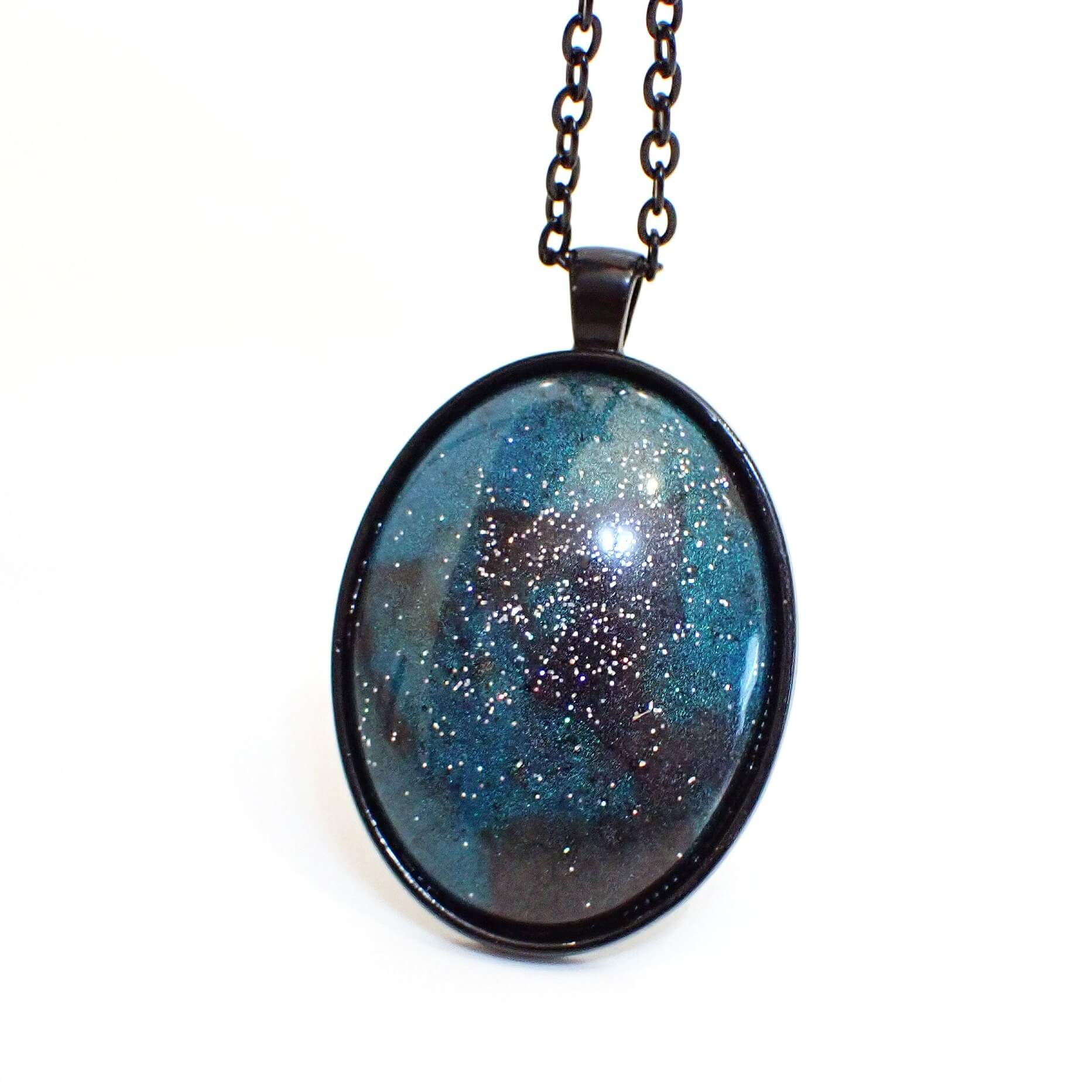 Enlarged front view of the handmade Goth pendant necklace. The metal is black coated in color on the chain and on the pendant setting. The pendant is a large domed oval with pearly dark gray and teal blue resin. There is silver holographic glitter embedded in the resin for tiny specks of sparkle and flashes of color as you move around in the light.