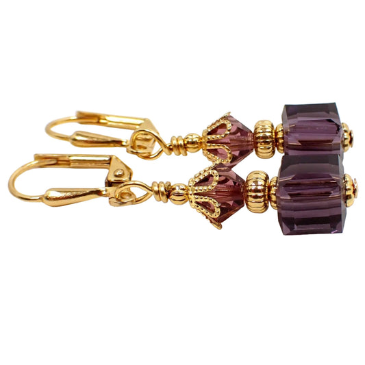 Side view of the small purple glass beaded earrings. The metal is gold plated in color. There are faceted glass crystal bicone beads at the top and small cube shaped glass beads at the bottom. The beads are purple in color.