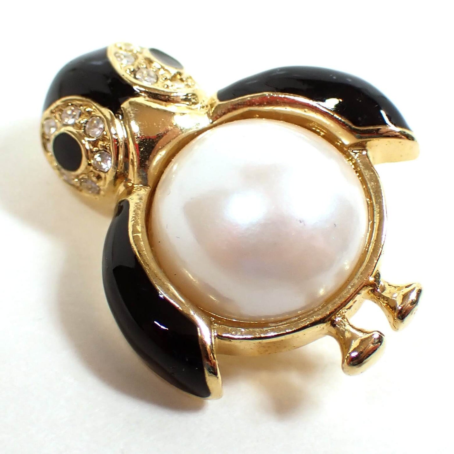 Enlarged front view of the small Marvella retro vintage brooch pin. It is shaped like a rounded penguin with black enameled head and wings and a plastic faux pearl belly. There are clear glass crystal rhinestones around the eyes. The metal is gold tone plated in color.