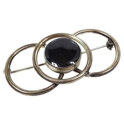 Front view of the retro vintage Taxco brooch pin. The sterling silver is darkened from age to a dark gray and even black in some areas. There are three open circles that overlap making up the brooch. The middle circle has a round area in the middle of it with black enamel. 