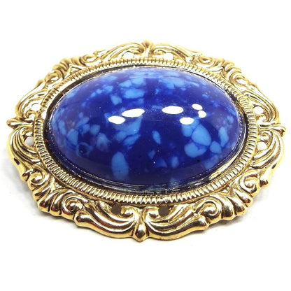 Front view of the retro vintage brooch pin. The metal is gold tone in color. It has an oval design with fancy style edge. The middle cab is a domed oval cab made of plastic that is dark blue with lighter blue speckles.
