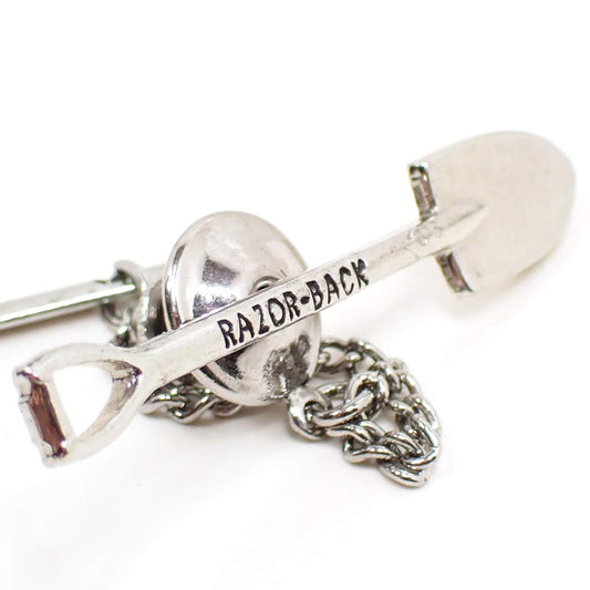 Enlarged front view of the retro vintage shovel tie tack. The metal is silver tone in color. It's shaped like a shovel and has the words Razor Back on the handle part in black.