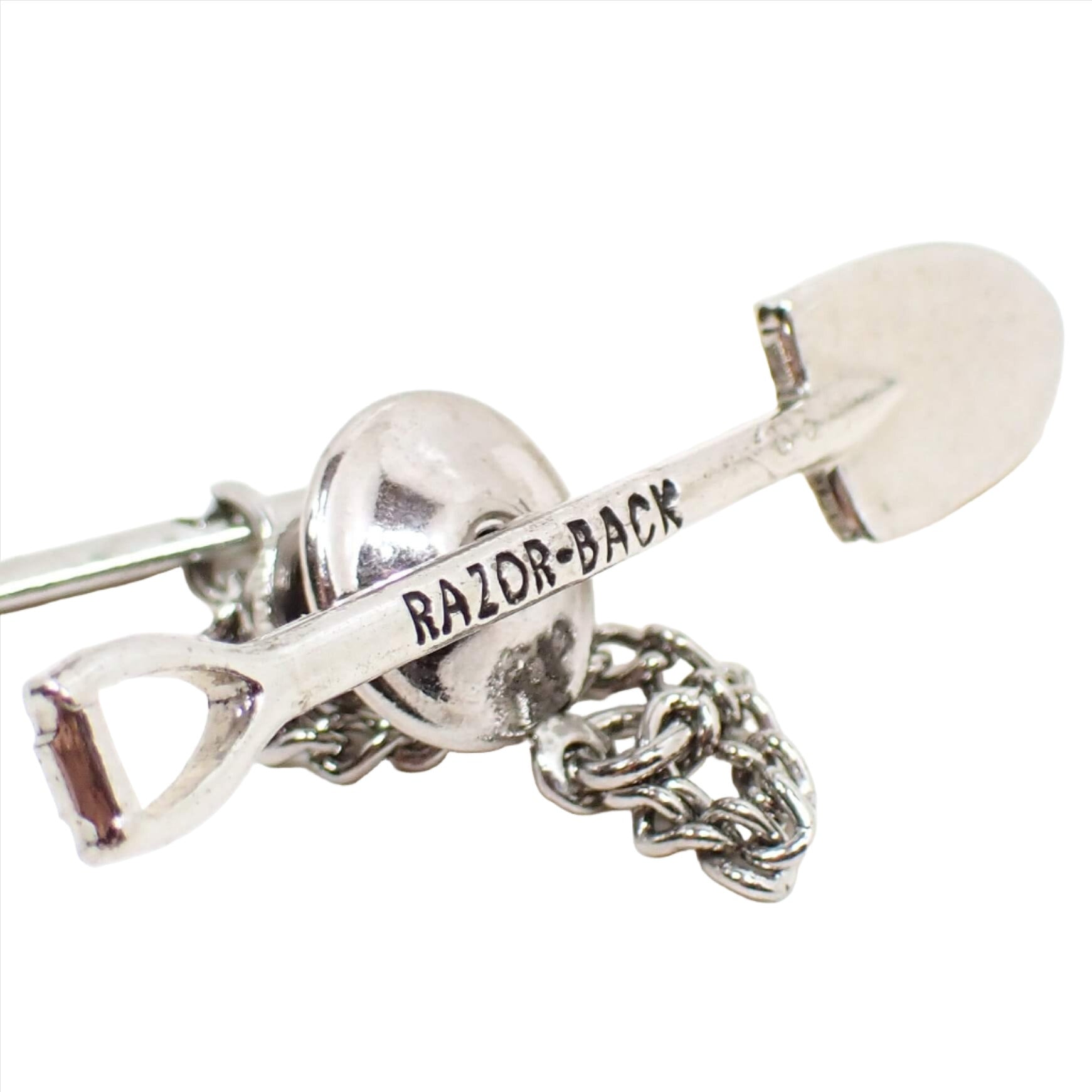 Enlarged front view of the retro vintage shovel tie tack. The metal is silver tone in color. It's shaped like a shovel and has the words Razor Back on the handle part in black.