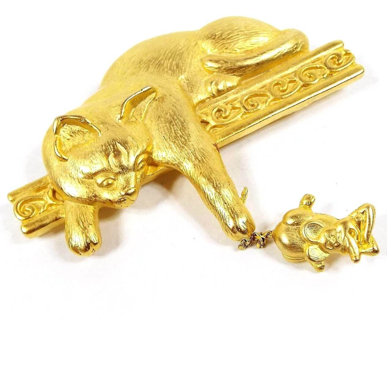 Front view of the retro vintage JJ cat and mouse brooch pin. The metal is mostly matte gold tone in color with some shiny edges. There is a cat laying across a fancy style bar with a curl design on it. The cat's paw is reaching down and holding the mouse by the tail. The mouse has a cartoon like appearance and has its arms folded in front of it like it's angry.