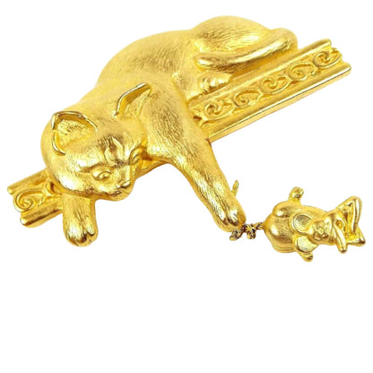 Front view of the retro vintage JJ cat and mouse brooch pin. The metal is mostly matte gold tone in color with some shiny edges. There is a cat laying across a fancy style bar with a curl design on it. The cat's paw is reaching down and holding the mouse by the tail. The mouse has a cartoon like appearance and has its arms folded in front of it like it's angry.