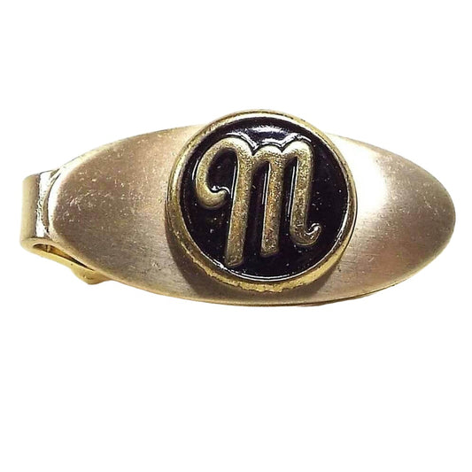Front view of the retro vintage initial tie clip. It is gold tone in color and has a small oval shape. There is a black enameled circle in the center with the letter M on it.