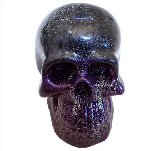 Front view of the handmade resin skull. The top part of the skull is pearly dark gray and black resin. The bottom half of the skull has areas with different shades of purple and dark gray. There is holographic silver glitter mixed in around the whole skull.