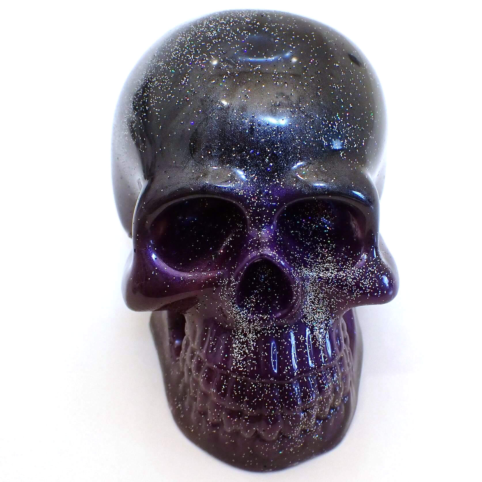 Front view of the handmade resin skull. The top part of the skull is pearly dark gray and black resin. The bottom half of the skull has areas with different shades of purple and dark gray. There is holographic silver glitter mixed in around the whole skull.