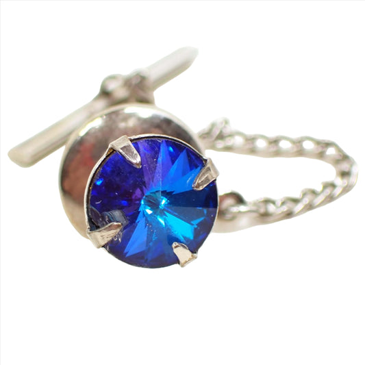 Front view of the Mid Century vintage rhinestone tie tack. The rhinestone is a round rivoli rhinestone that is faceted and comes slightly outward to a pointed middle. It has varying shades of blue from deep sea to aqua blue as you move around. The rhinestone is set in a prong setting and is silver tone in color. The back clutch is rounded with a chain that has a small bar at the end.