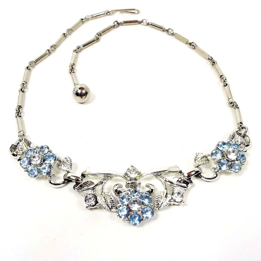 Front view of the Mid Century vintage rhinestone floral necklace. The metal is silver tone in color. It has a link design with bar links down to three flower links. The middle link is the largest. The flower petals are light blue in color and the rest of the rhinestones are clear. 