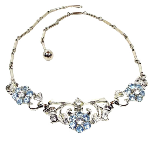 Front view of the Mid Century vintage rhinestone floral necklace. The metal is silver tone in color. It has a link design with bar links down to three flower links. The middle link is the largest. The flower petals are light blue in color and the rest of the rhinestones are clear. 