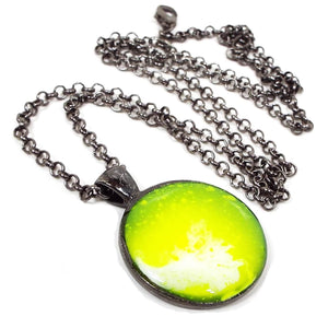 Front view of the handmade resin pendant. The metal is gunmetal plated dark gray. There is a rolo chain with a lobster claw clasp at the end. The round pendant has white and neon yellow swirls and spots. 