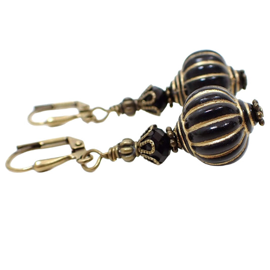 Side view of the handmade lantern drop earrings. The metal is antiqued brass in color. There are black faceted glass crystal beads at the top and black acrylic lantern shape beads at the bottom. The bottom beads have antiqued gold stripes painted on them.