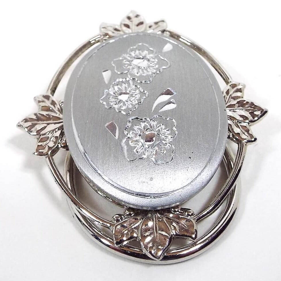 Front view of the retro vintage Jeri-Lou floral scarf clip. The metal is silver tone in color. The frame around the edge has an open wire style design with leaves at the top, bottom, and sides. The large oval cab in the middle is a matte silver color and has an etched flower design with three flowers and an etched line around the edge.