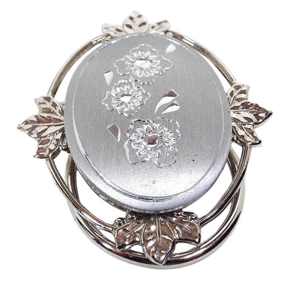 Front view of the retro vintage Jeri-Lou floral scarf clip. The metal is silver tone in color. The frame around the edge has an open wire style design with leaves at the top, bottom, and sides. The large oval cab in the middle is a matte silver color and has an etched flower design with three flowers and an etched line around the edge.